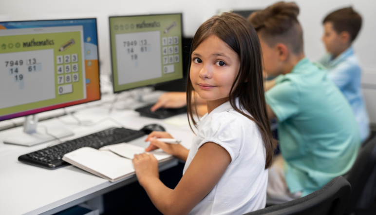 Young girl looking at camera while using a computer in a classroom with maths program on screen.